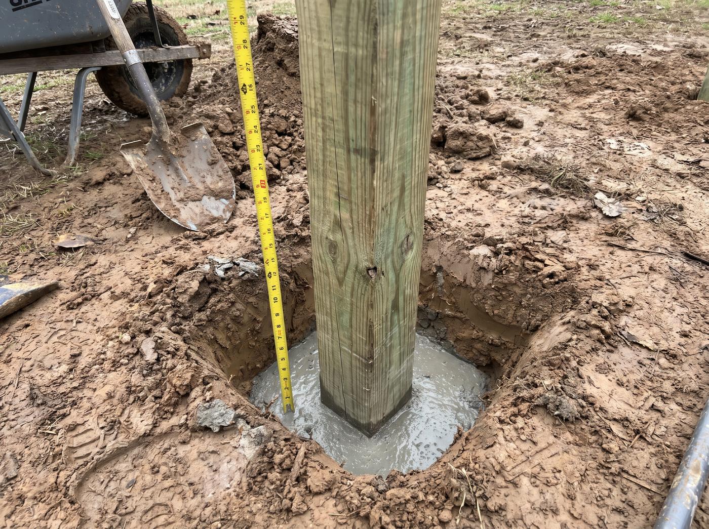 A wooden fence post set in a concrete footing in clay soil, with a tape measure in the hole